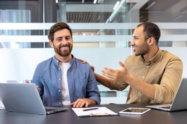 İki farklı erkek meslektaşı, dizüstü bilgisayarlar ve diğer dijital cihazlar kullanarak modern ofis ortamında gülümsüyor ve işbirliği yapıyorlar..