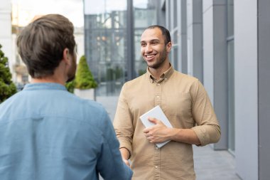 Modern bir binanın önünde el sıkışan iki işadamı. Bir adam tableti tutuyor ve gülümsüyor..