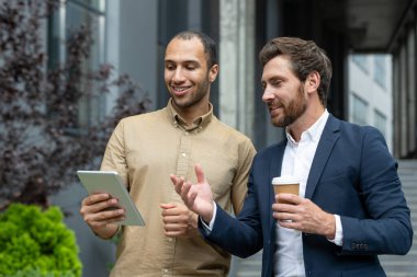 İki işadamı bir ofis binasının yakınındaki tableti inceliyor, doğal ifadeler ve gülümsemelerle kahve tartışıyor ve içiyorlar..
