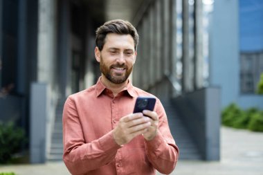 Sakallı bir adam modern bir binanın dışında telefonuna bakarken gülümsüyor. Düğmeli bir gömlek giyiyor..