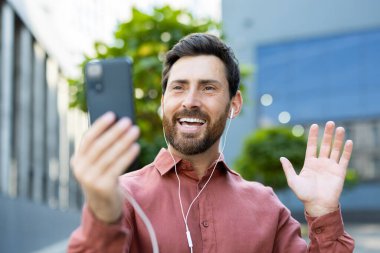 Sakallı bir adam akıllı telefonuyla bir video görüşmesinde gülümsüyor ve el sallıyor..