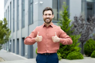 Dışarıda gülümseyen bir adam baş parmağını kaldırıyor. Pembe bir gömlek ve koyu mavi pantolon giyiyor..