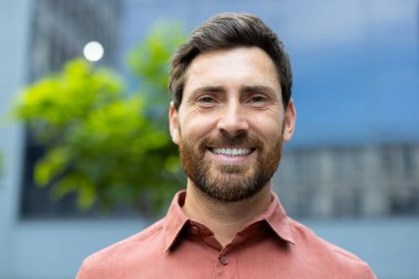 Yakışıklı bir adam ışıl ışıl gülümser, gözleri parıldar. Dışarıda, doğrudan kameraya bakıyor. Sıcaklık ve yakınlık yayıyor..