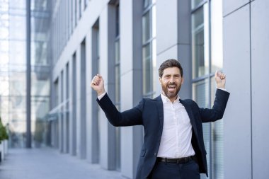 Başarılı bir işadamı başarısını bir ofis binasının önünde sevinçle kollarını kaldırarak kutlar..