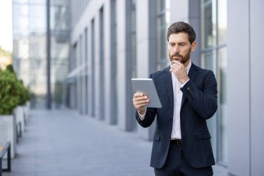 Takım elbiseli odaklanmış bir işadamı modern bir ofis binasının önünde tablet kullanır..