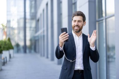 Takım elbiseli gülümseyen bir işadamı, akıllı telefonuyla yaptığı bir video görüşmesi sırasında modern bir binanın yanında duruyor..