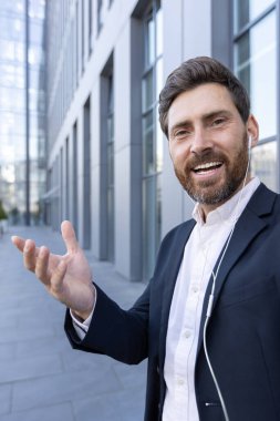 Gülümseyen bir iş adamı, modern bir binanın önünde görüntülü bir aramada, takım elbise giyiyor ve kulaklık takıyor, eliyle sunum yapıyor.