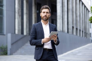 Takım elbiseli bir işadamı elinde tabletle arka planda bir binayla dışarıda duruyor..