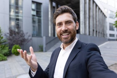 Videodaki bir iş adamı, binanın önünde gülümsüyor ve el kol hareketi yapıyor.