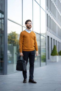 Bir işadamı, modern bir binanın önünde, elinde bir evrak çantasıyla, kendinden emin bir şekilde duruyor..