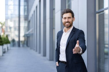 Takım elbiseli bir işadamı modern bir ofis binasının önünde el sıkışarak gülümsüyor..