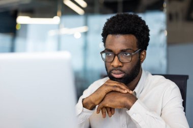 Odaklanmış, gözlüklü bir adam dikkatle bilgisayar ekranına bakar, iş yerinde derin düşüncelere dalmıştır..