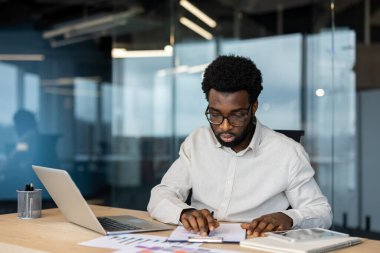Belgeler üzerinde çalışan ve dizüstü bilgisayar kullanan, modern bir ofiste çalışan Afrikalı bir iş adamı..