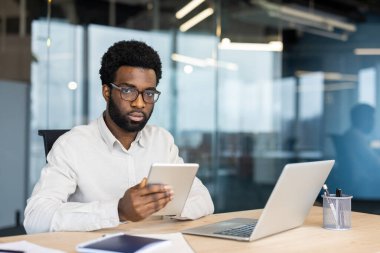 Gözlüklü bir Afro-Amerikalı ofis masasında tablet ve dizüstü bilgisayarla çalışıyor. Kameraya bakıyor..