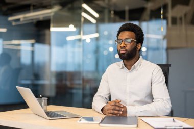 Gözlüklü düşünceli bir Afro-Amerikalı ofiste bir masada dizüstü bilgisayar ve not defteriyle oturuyor..