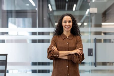 Gülümseyen bir kadın, güven içinde bir ofis ortamında, kollarını kavuşturmuş, profesyonellik ve yakınlık saçıyor..