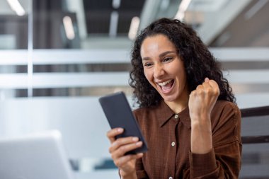 Neşeli bir kadın, mutlu haberi telefonundan kutluyor. Yumruğu zafer dolu bir gülümsemeyle yükseliyor..