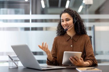 Kulaklık takan gülümseyen bir kadın, elinde tabletle bir ofis ortamında video görüşmesi yapıyor..