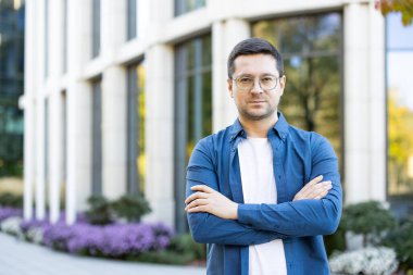 Bir adam, modern bir binanın önünde kollarını kavuşturmuş, profesyonel bir aura yayıyor..