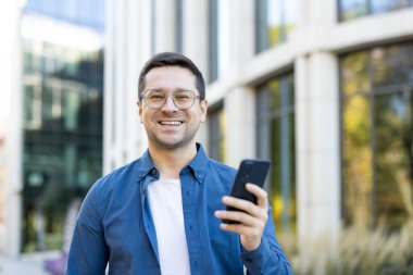 Güneşli bir günde modern bir binanın önünde telefonu tutan gülümseyen bir adam..