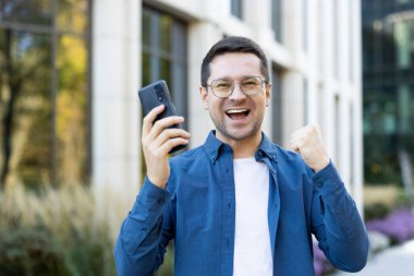 Elinde telefon tutan bir adam, bir yumrukla zaferi kutluyor. Dışarıda, bir binanın önünde..