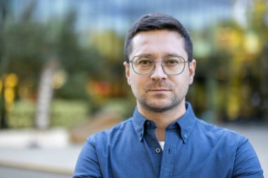 Gözlüklü ve mavi gömlekli bir adam kameraya ciddi bir ifadeyle bakıyor..