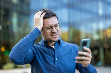 Gözlüklü, hayal kırıklığına uğramış bir adam telefonuna bakıyor, inançsızlıkla kafasına dokunuyor. Sahne modern bir binanın yanında..