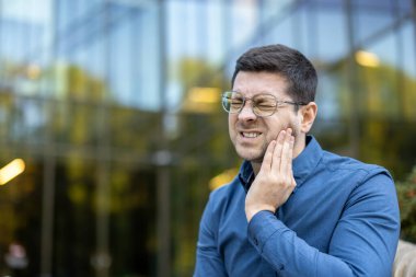 Ağrı çeken bir adam, muhtemelen diş ağrısı, yanağına bir rahatsızlık ifadesi ile dokunuyor. Gözlük takıyor..