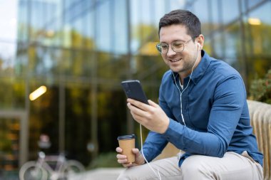 Bir adam dışarıda oturmuş, müzik dinlerken telefonunu kullanıyor. Bir kahve fincanı tutuyor ve modern bir binanın önünde gülümsüyor..