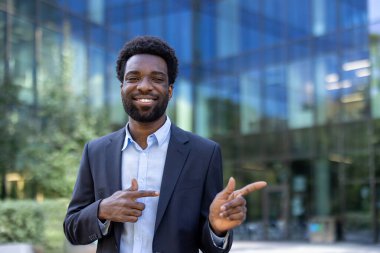 Gülümseyen bir işadamı modern bir ofis binasının önünde dururken parmaklarını sağa çevirir..