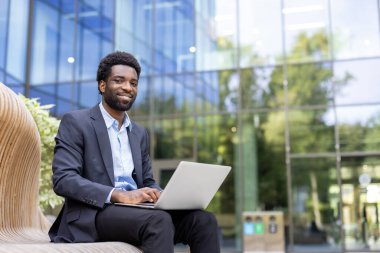 Takım elbiseli, gülümseyen Afro-Amerikalı bir adam dışarıda bir bankta otururken bilgisayarında çalışıyor.