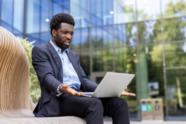 Takım elbiseli sinirli bir işadamı dizüstü bilgisayarına bakarken tepki veriyor. Açık havada, modern bir binanın yanında..