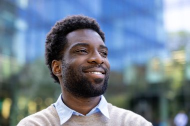 Gülümseyen bir adam uzaklara bakar, muhtemelen şehir ortamında, pozitiflik ve güven yayar..