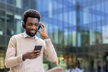 Afro-Amerikalı bir adam kulaklıkla müzik dinlerken gülümsüyor, telefonunu modern bir binanın dışında tutuyor..