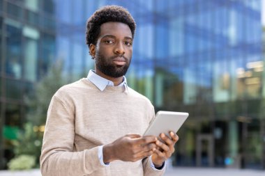 Hafif kazaklı Afro-Amerikan bir adam, elinde tablet, bir ofis binasının önünde poz veriyor, kameraya bakıyor..