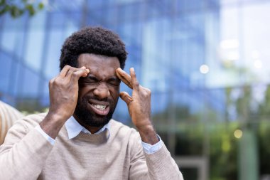 Baş ağrısı çeken bir adam, parmaklarını şakaklarına bastırıyor açık havada gözleri kapalı..