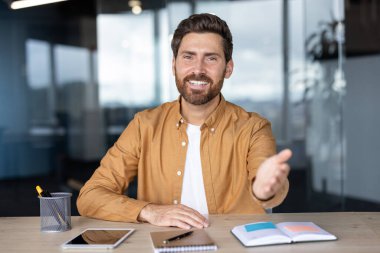 Gülümseyen bir işadamı, ofis ortamında el sıkışmaya hazır bir şekilde masasında hoş geldin jesti yapar..