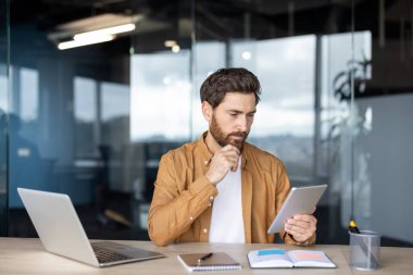 Sakallı düşünceli bir adam ofis ortamında bir tablete bakıyor, muhtemelen verileri analiz ediyor ya da bir proje üzerinde çalışıyor..