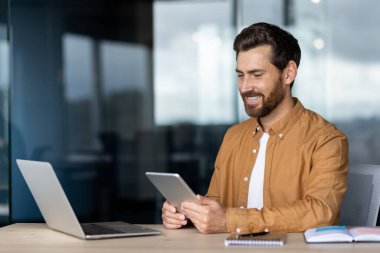 Gülümseyen bir adam masada oturuyor, elinde tablet, yanında dizüstü bilgisayarla, ofisin içinde..