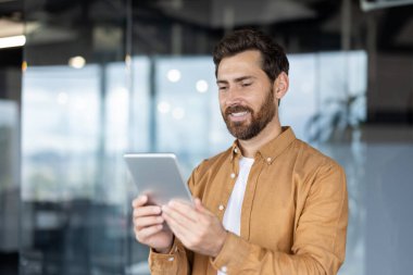Gülümseyen bir işadamı modern ofis ortamında bir tablet tutuyor, bu da katılımını ve teknoloji kullanımını gösteriyor.