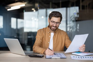 Gözlüklü neşeli bir işadamı, analiz için dizüstü bilgisayar ve hesap makinesi kullanarak ofisindeki mali belgeleri inceliyor..