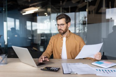 Ofis masasında bir işadamı, dizüstü bilgisayarla çalışıyor, elinde kağıtlar tutuyor, verileri analiz ediyor ve gözlük takıyor..