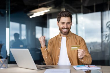 Adamın biri dizüstü bilgisayarın yanında kredi kartıyla kutlama yapıyor. Heyecan gösteriyor. Sahnede çevrimiçi alışveriş veya başarılı bir mali işlem tasvir ediliyor.