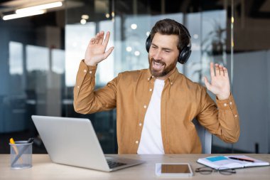 Kulaklık takan bir adam muhtemelen video görüşmesi sırasında ya da müzik dinlerken dizüstü bilgisayarının önünde el kol hareketi yapıyor..