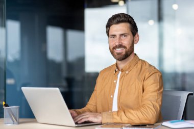 Gülümseyen bir işadamı, modern ofis ortamında dizüstü bilgisayarda çalışarak profesyonelliği ve üretkenliği aktarıyor..