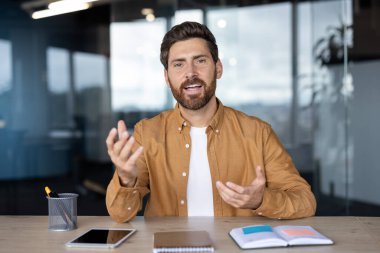 Videodaki bir adam konuşurken heyecanla el kol hareketi yapar, muhtemelen ofis ortamındaki bir masada..