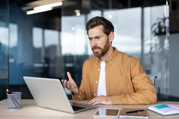 A concerned businessman in a bright orange shirt gestures while looking at his laptop during a video call.
