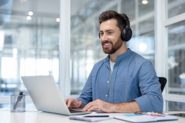 Kulaklık takan gülümseyen bir adam modern ofis ortamında dizüstü bilgisayarda çalışır, mutlu görünür..