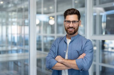 Gözlüklü, gülümseyen bir işadamı, bir ofiste, kollarını kavuşturmuş, başarının ve ulaşılabilirliğinin bir resmi olarak duruyor..