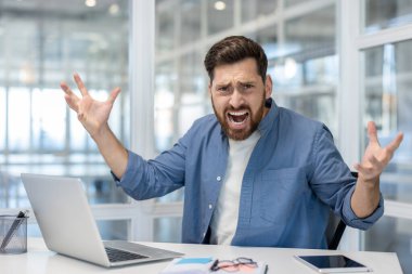 Bir adam görünür bir şekilde üzgün ve bağırıyor, kolları havada, ofisinde masasında..